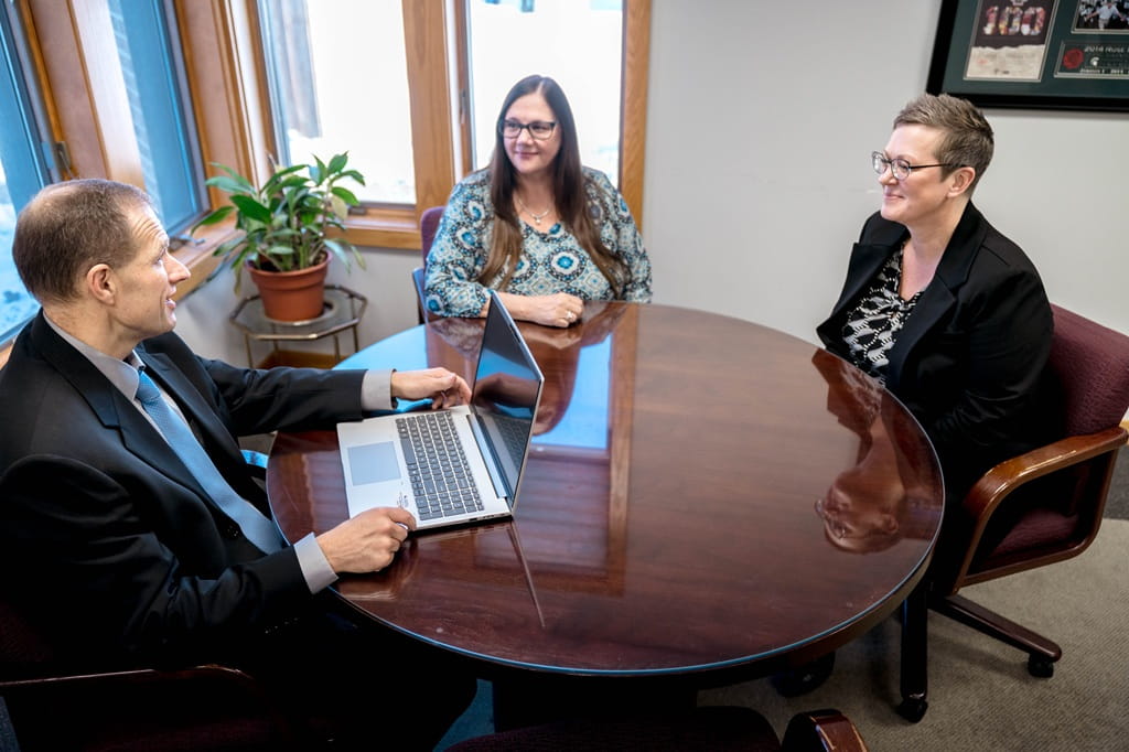 A male financial advisor with a laptop talks with two female clients at a round meeting table.