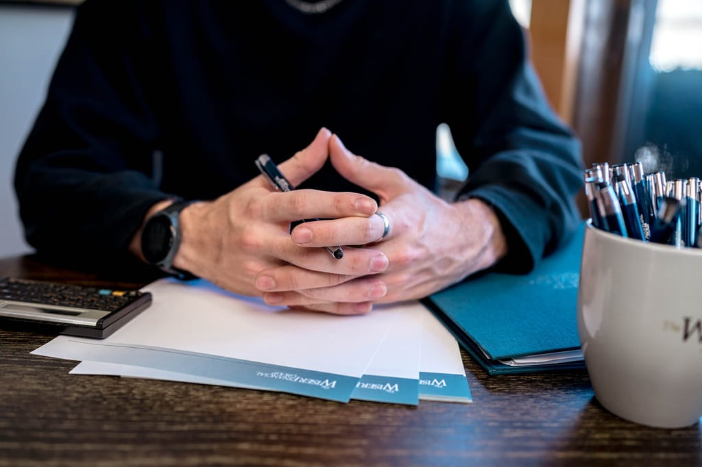 A financial advisors hands interlocked over paperwork with a coffee cup full of pens on the desk next to them.
