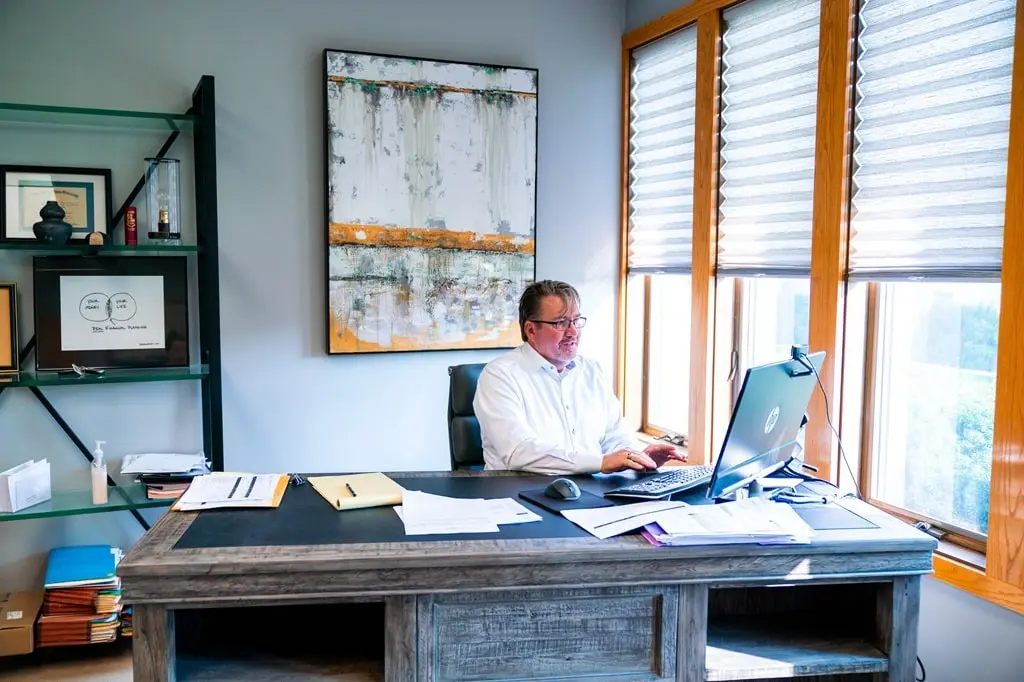 A man with a painting behind him sits at a work desk with papers, a laptop, and a mouse, typing at the laptop.