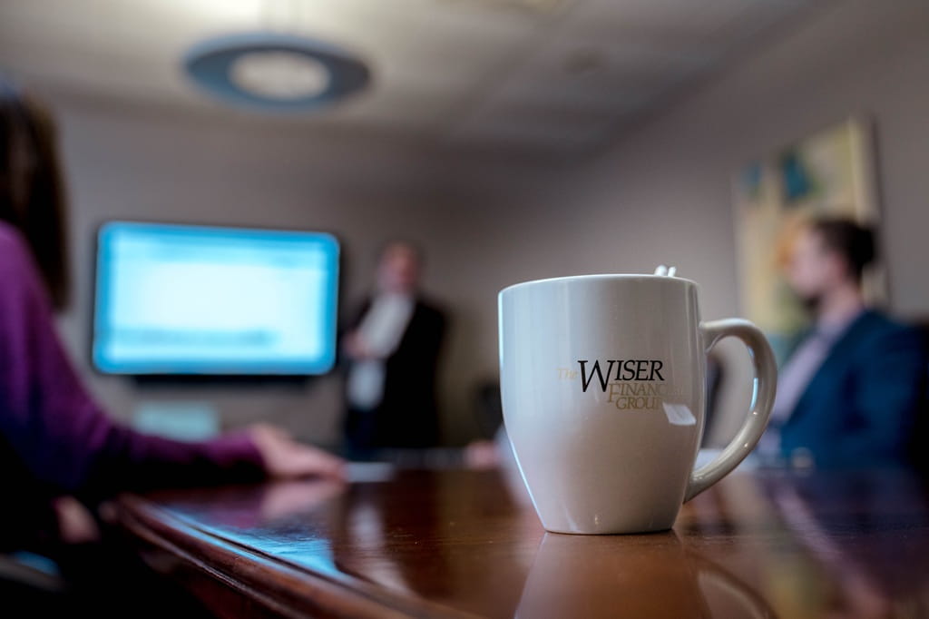 A focused photo of a Wiser Financial Group coffee mug, with a few team members blurred in the background as they sit in a team meeting looking at a presentation.